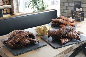 Auf einem Holztisch stehen drei Schieferplatten mit aufgetürmten Spareribs, etwa 5-6 Ribs pro Platte. In der Mitte des Tisches steht ein Brotkorb. Im Hintergrund die Getränkekarte des Restaurants Huba in Neuburg.
